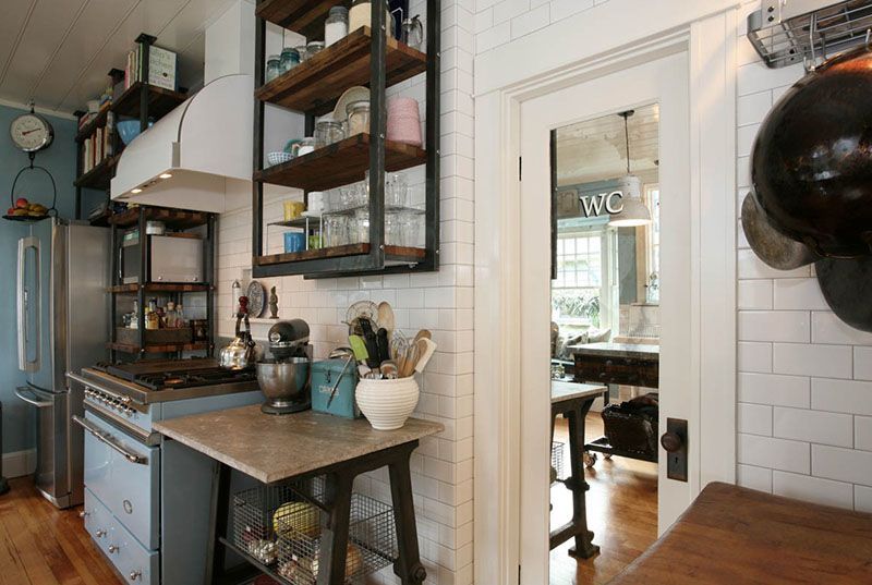 Small kitchen with blue cabinets, open shelves, and a white-tiled wall opening into a dining room.