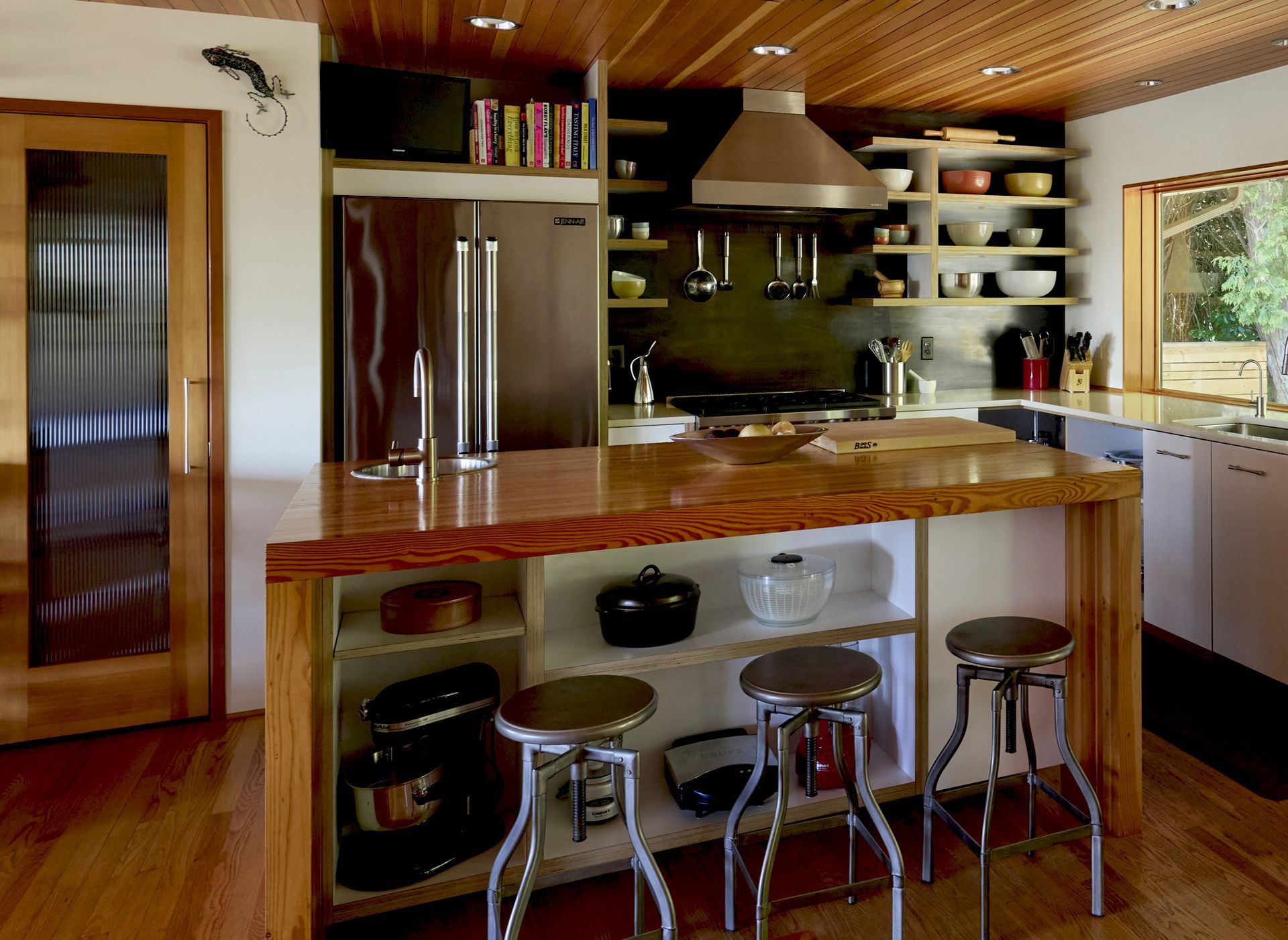 Wide view of the kitchen looking at the range wall past the substantial fir-clad island with open cubbies below and a trio of industrial stools. The fir-paneled ceiling and oak floors pair beautifully with white walls and stainless steel.
