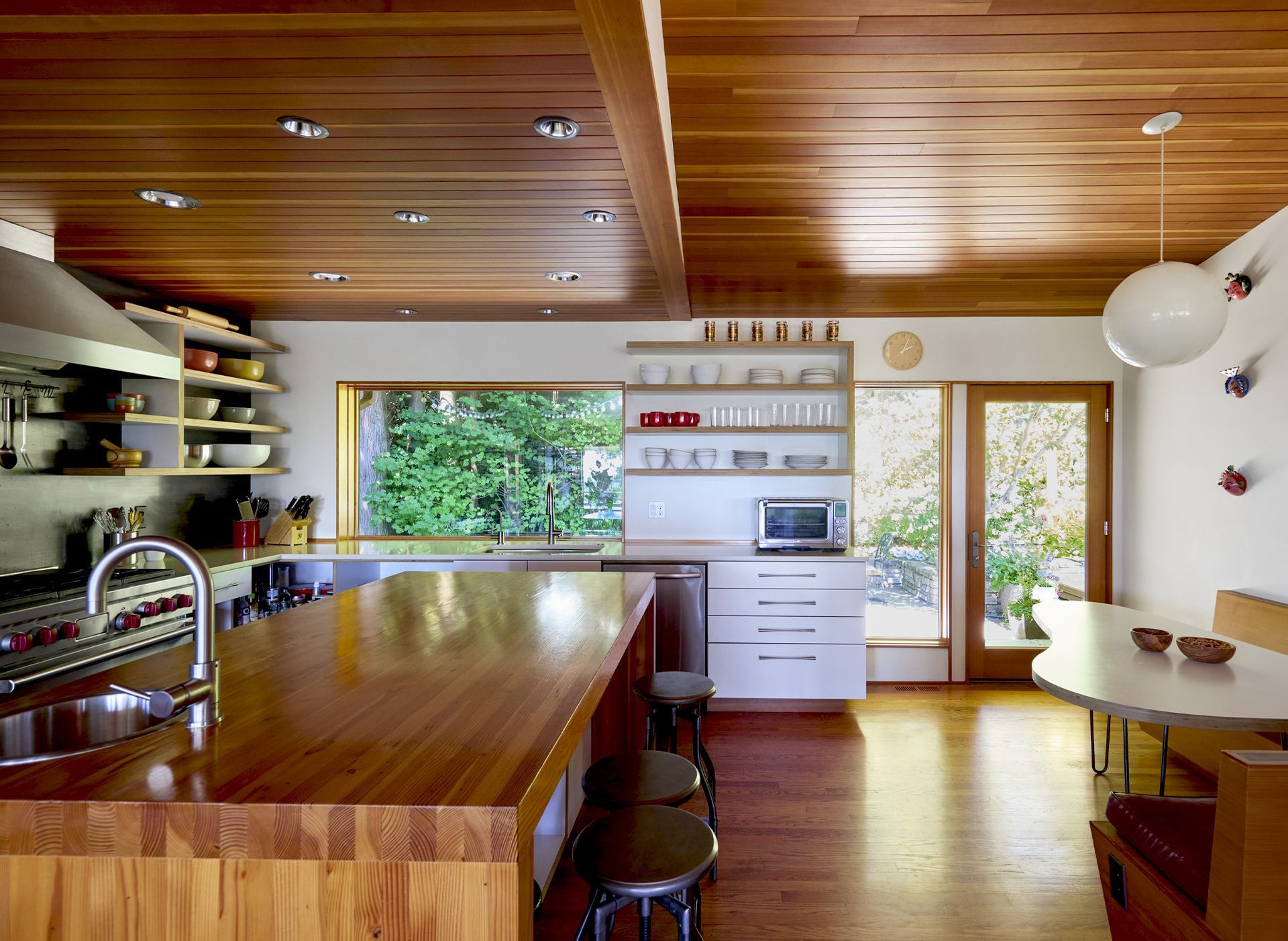 View of the East wall of the kitchen, with the sink, dishwasher, lower cabinets and open apple ply upper shelves, two picture windows and full lite door. A generous fir-clad island sits at the center of the room between the range wall and banquette.