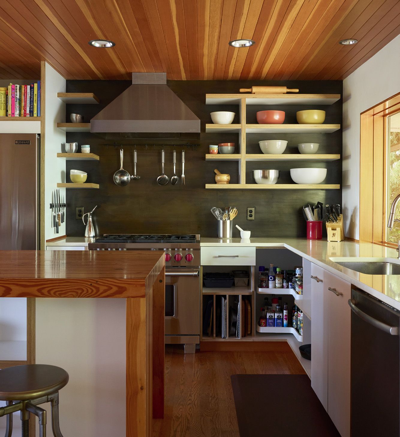 The North wall of the kitchen is home to the refrigerator and pro style range. A blackened steel backsplash contrasts with broad melamine and apple ply open shelves that display favorite utensils and colorful bowls.