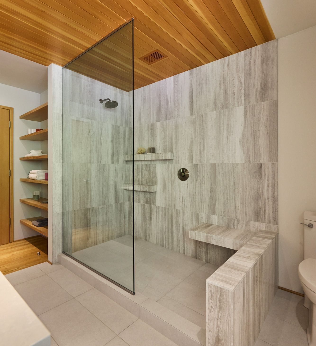 This spacious shower is clean and modern in limestone-inspired porcelain tile set vertically. It has a low curb, fixed glass panel, built-in bench and shelf, and easily fits two bathers.
