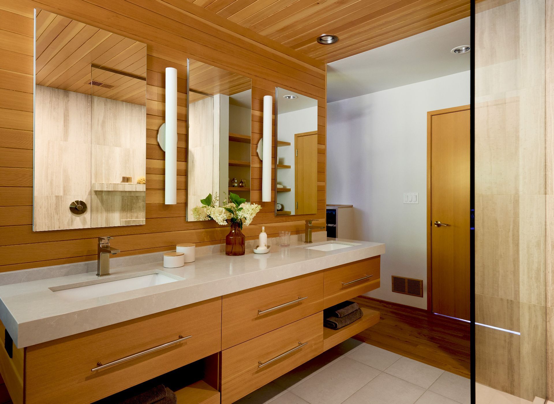 In the main bath, the centrally-located, vanity wall divides the room into bathing and closet areas with circulation all around it. The ceiling and backsplash wall at the floating double vanity are clad in warm-hued fir, cohesive with the cabinetry and harmonious with the warm white walls.