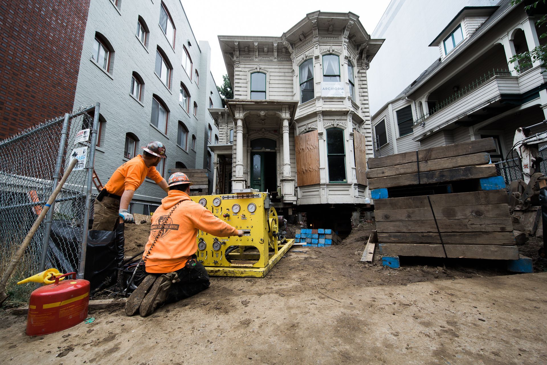 Construction workers in orange jackets repair a damaged building beside a muddy lot and heavy equipment.