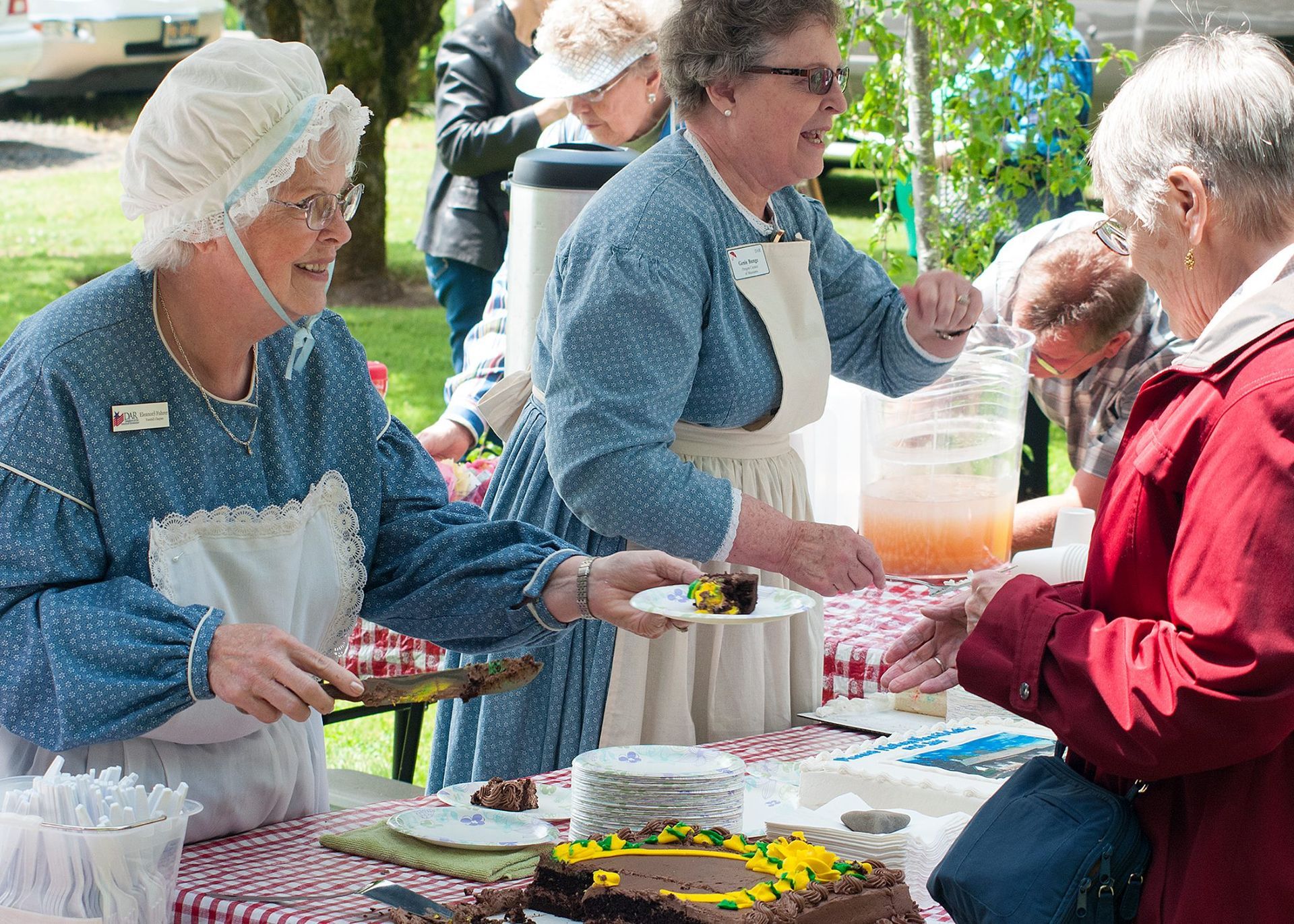 People serving food at an outdoor table with trays and a drink dispenser
