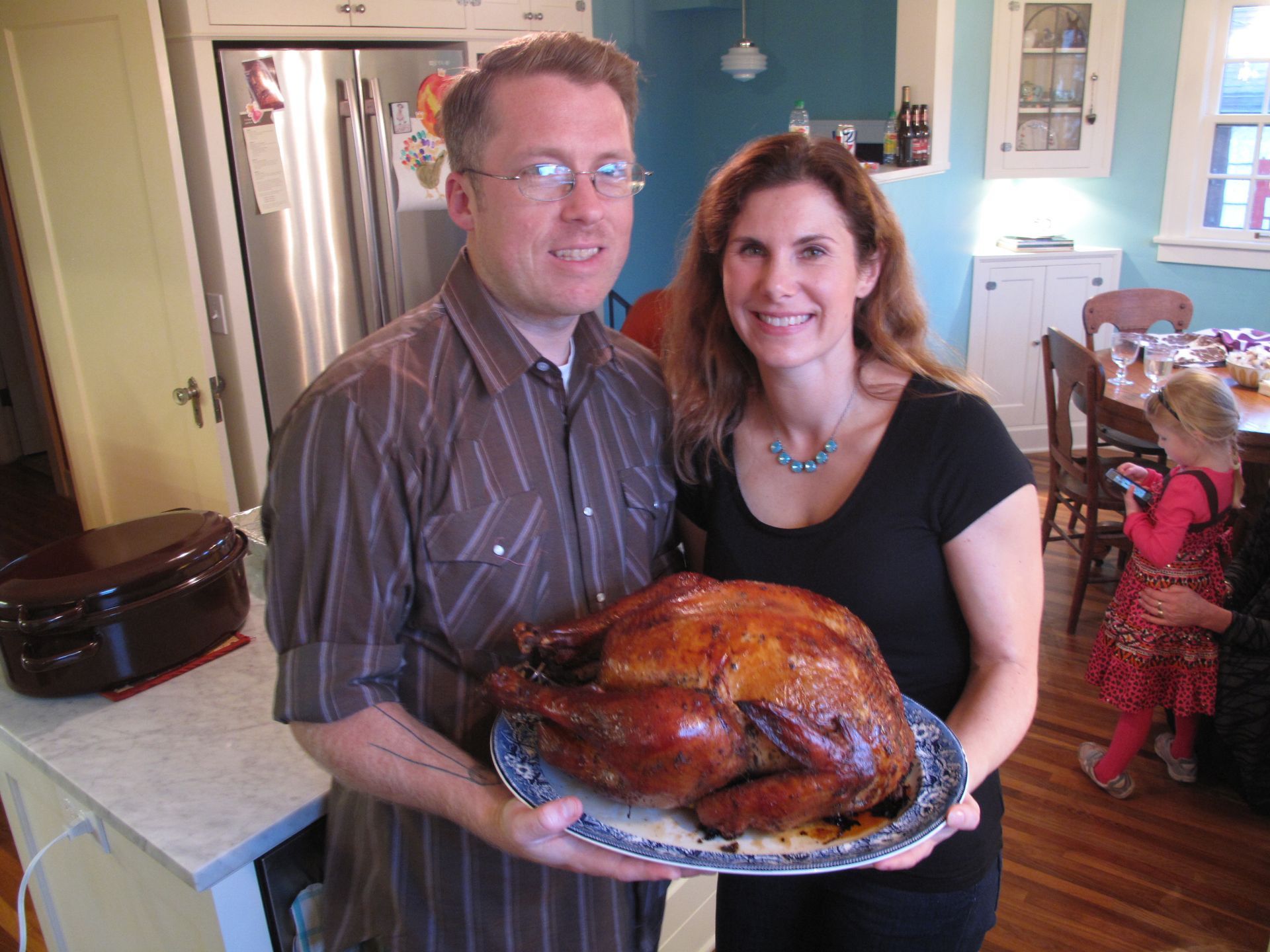 Couple in a kitchen holding a roasted turkey on a platter, with a child in the background.