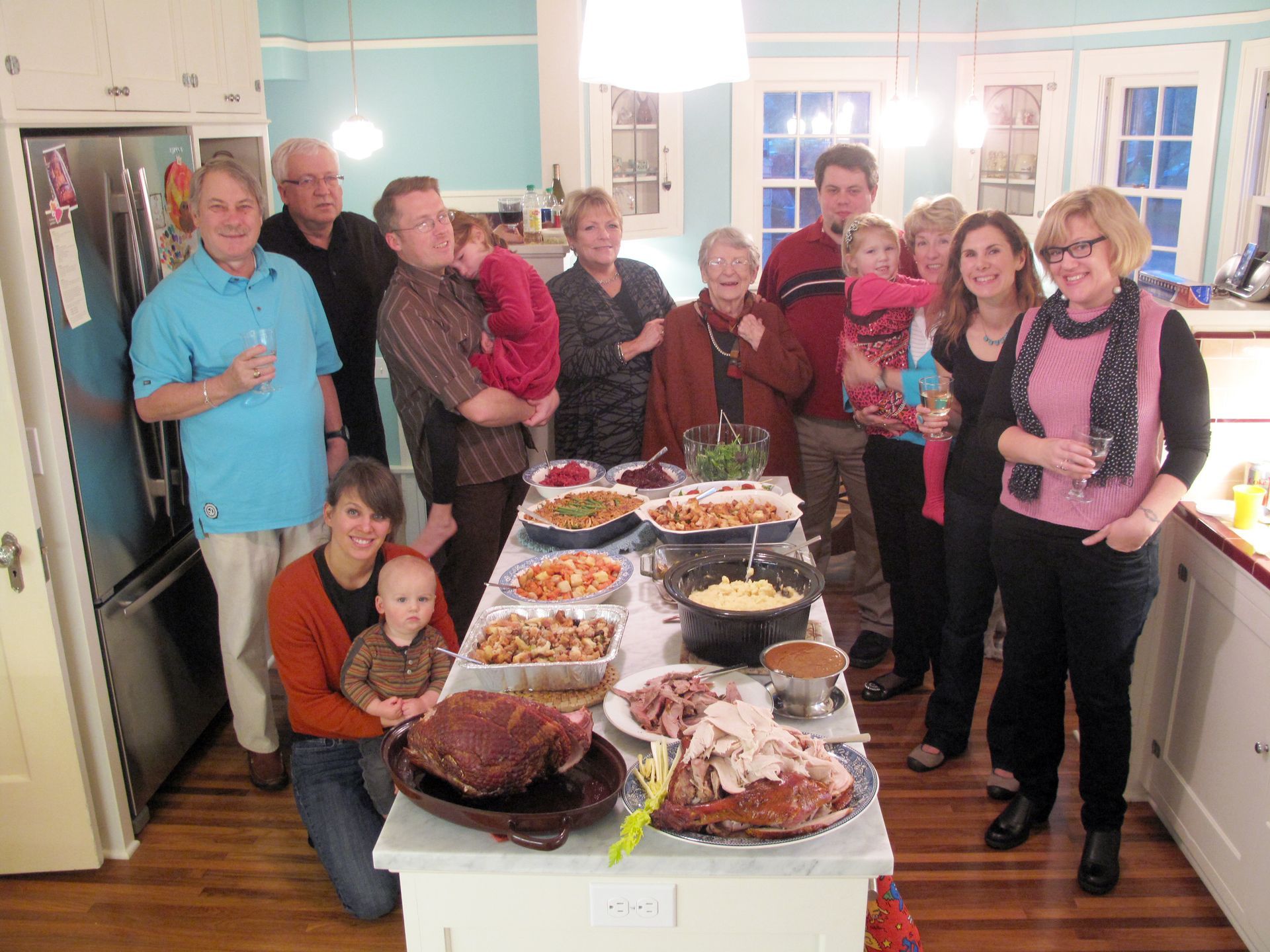 Group gathered around a table of food in a bright kitchen, smiling and holding drinks.