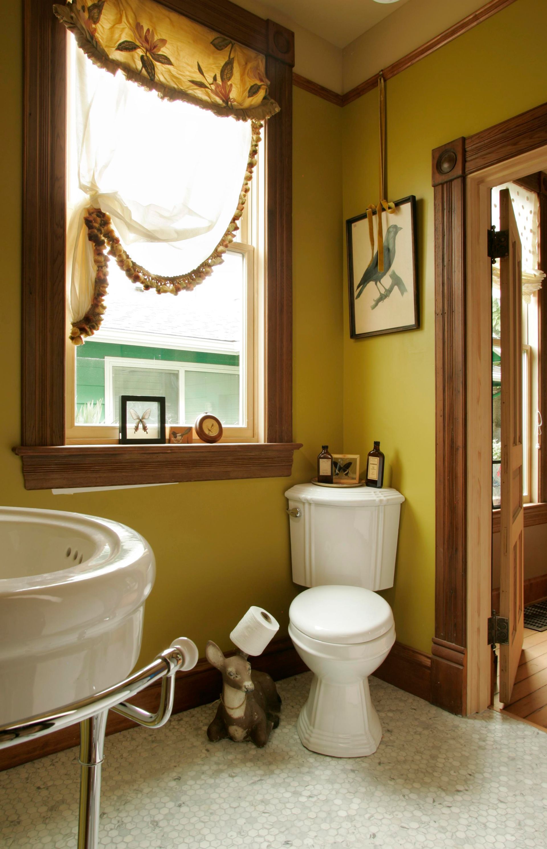 Small bathroom with toilet, sink, window, and brown trim; dog standing on the floor by the toilet.