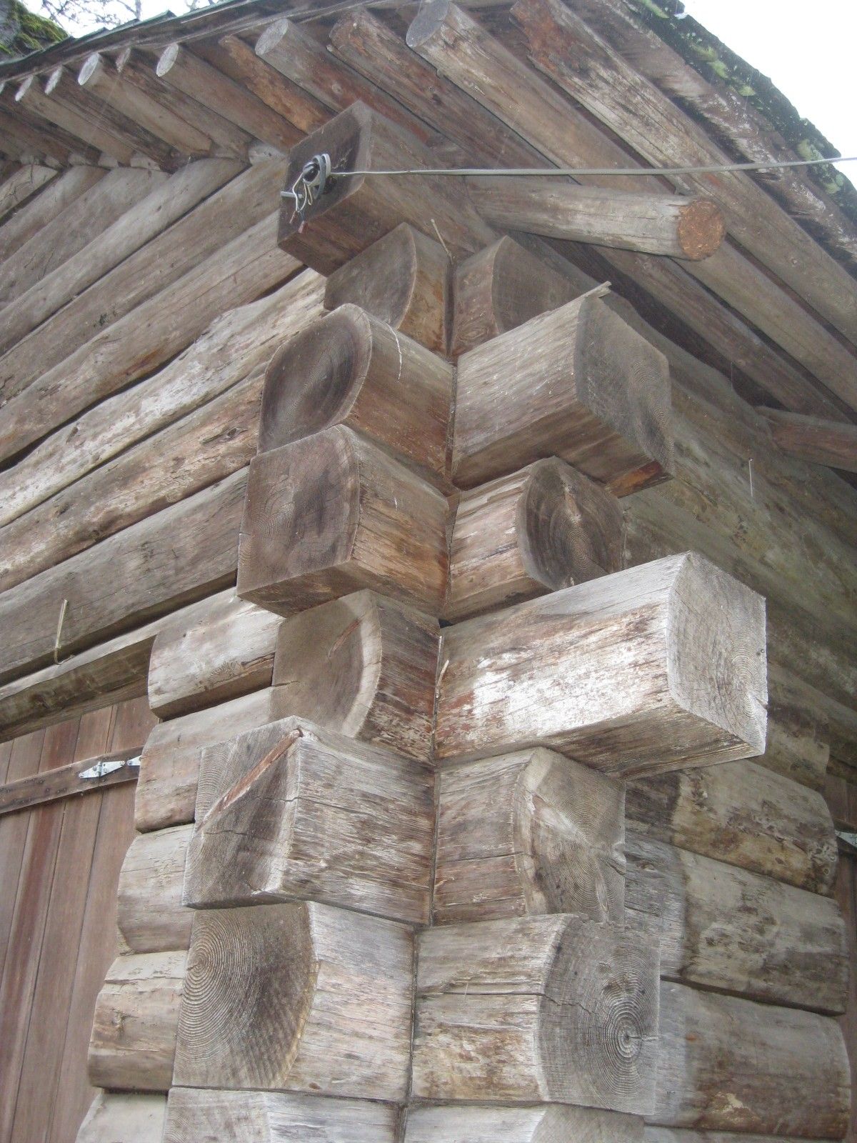 Weathered log corner of a wooden cabin with rough-hewn beams and slanted roof boards.