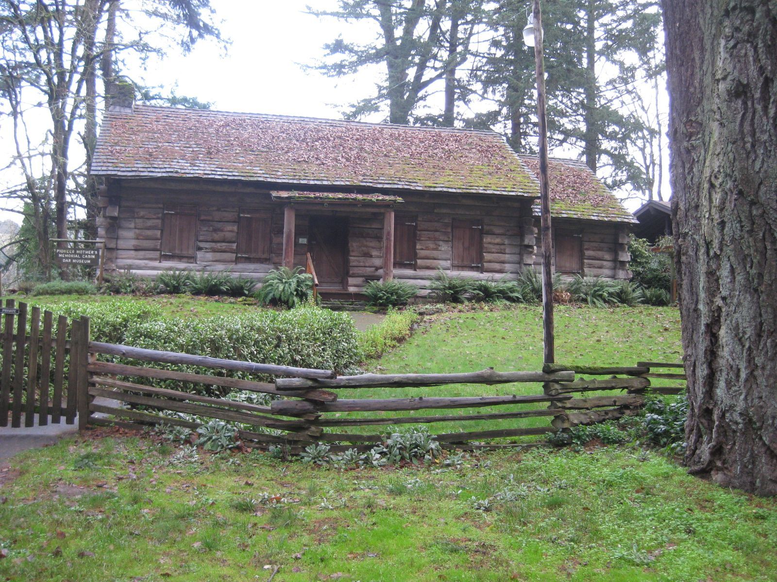 Old wooden cabin with a red roof behind a rustic fence in a grassy yard.
