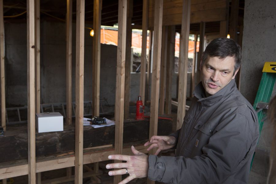 Man in a dark jacket gesturing inside an unfinished room with exposed wooden studs