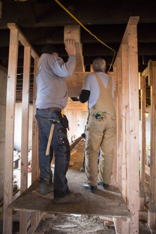 Two workers in a wood-framed house under construction, standing on a platform and measuring overhead.
