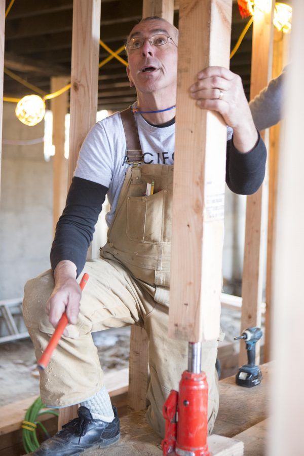 Construction worker kneeling beside a stud wall, using a red tool in an unfinished room.