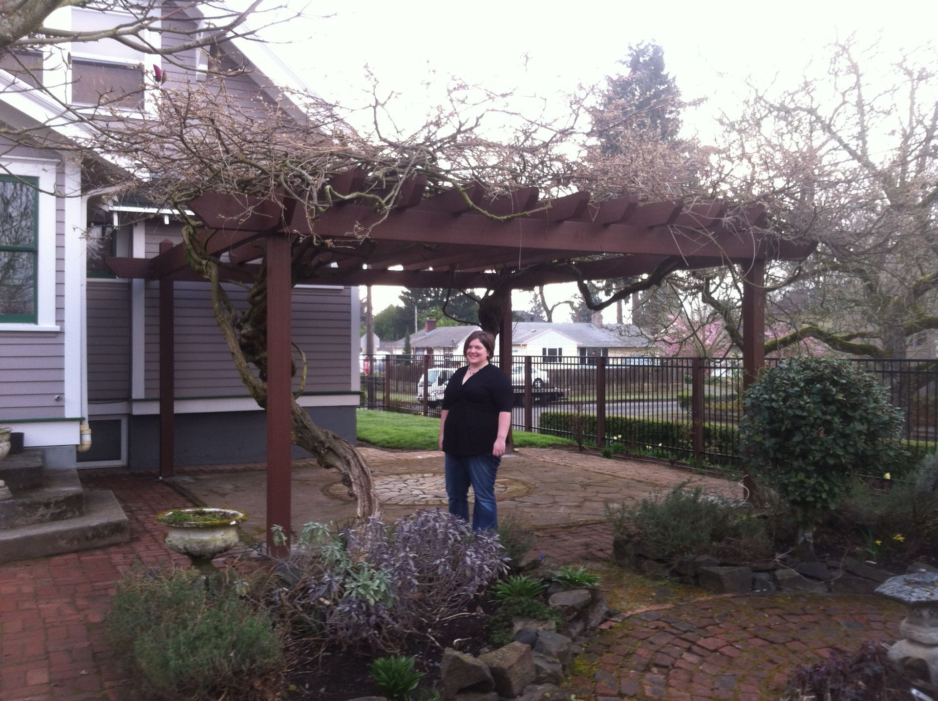 Person standing in a garden beside a vine-covered pergola near a house
