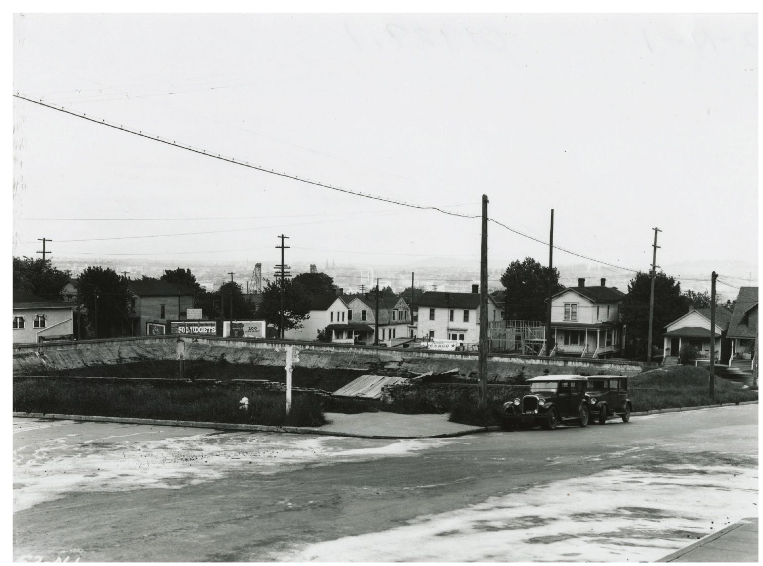 Black-and-white photo of a street with houses, utility poles, and parked cars behind a low wall