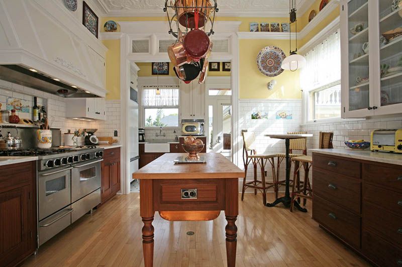 Bright kitchen with wood island, stainless stove, white cabinets, and sunlight through large windows.