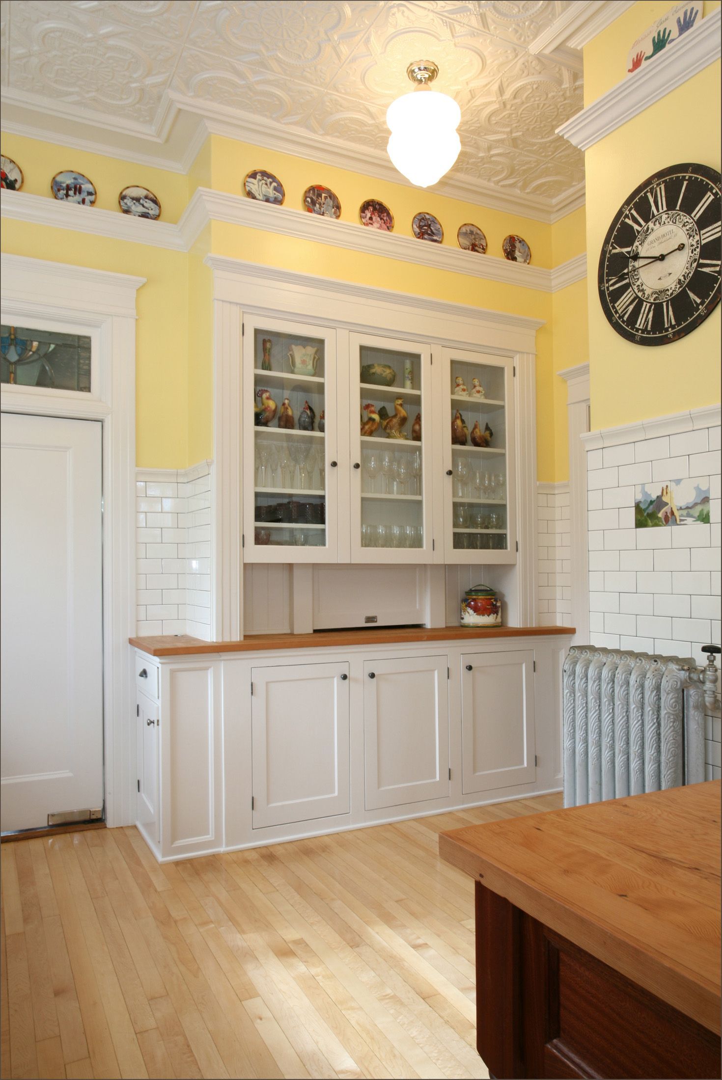Bright yellow kitchen with white cabinets, glass-front hutch, wooden floor, and a large black wall clock.