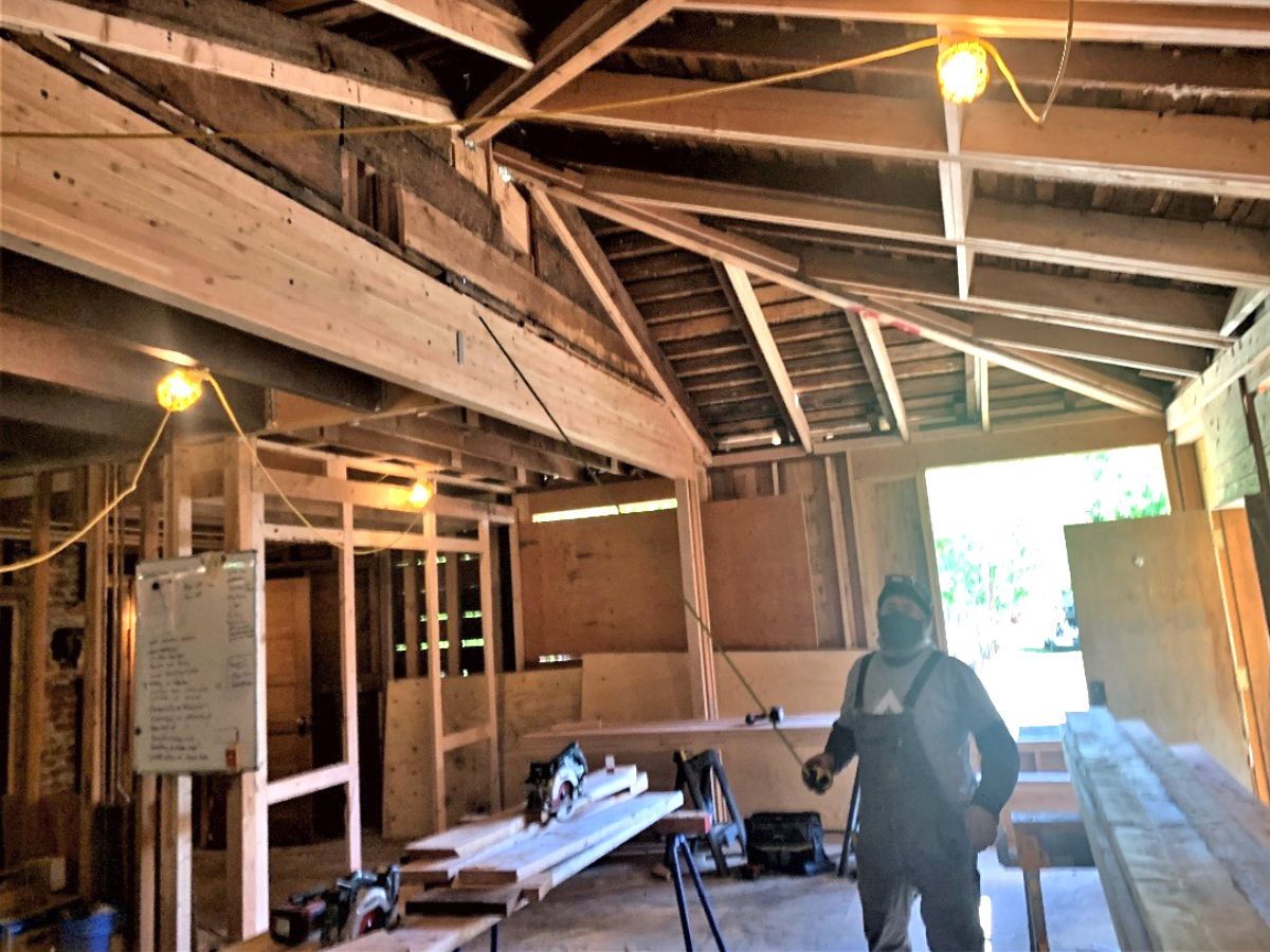 A person in a workshop surrounded by lumber and construction tools. Overhead beams, open walls and ceiling.