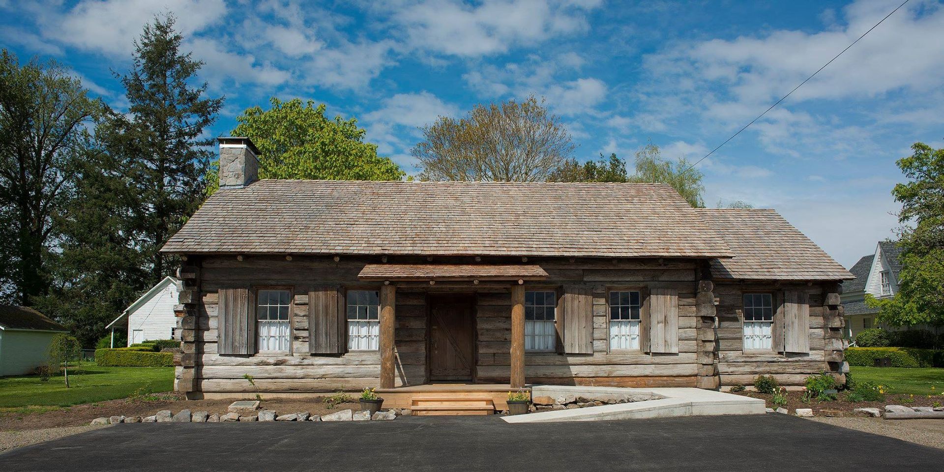 Rustic log cabin with a wooden porch, stone chimney, and trees under a blue sky