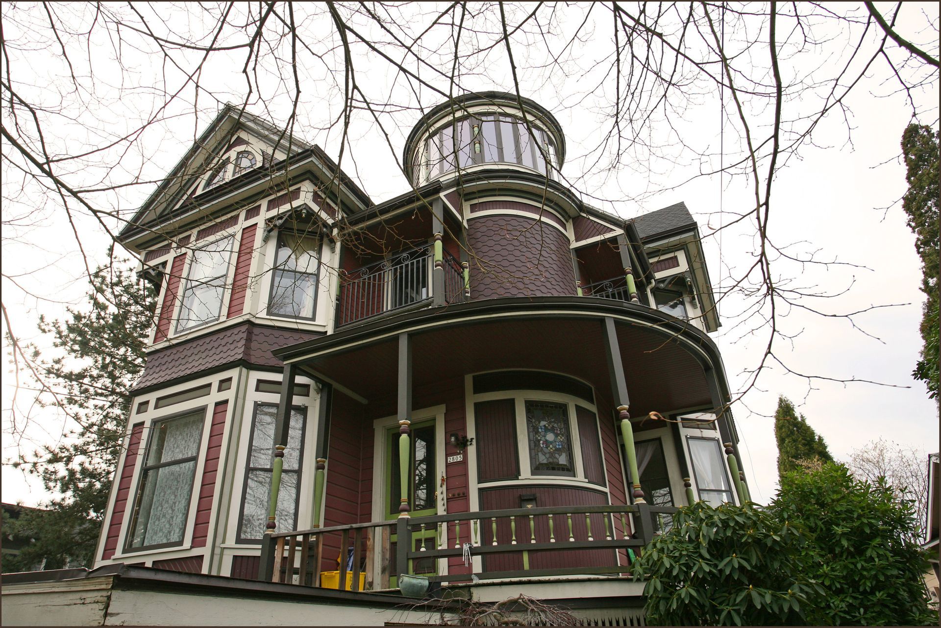 Victorian house with a wraparound porch and turret, shown among bare tree branches.