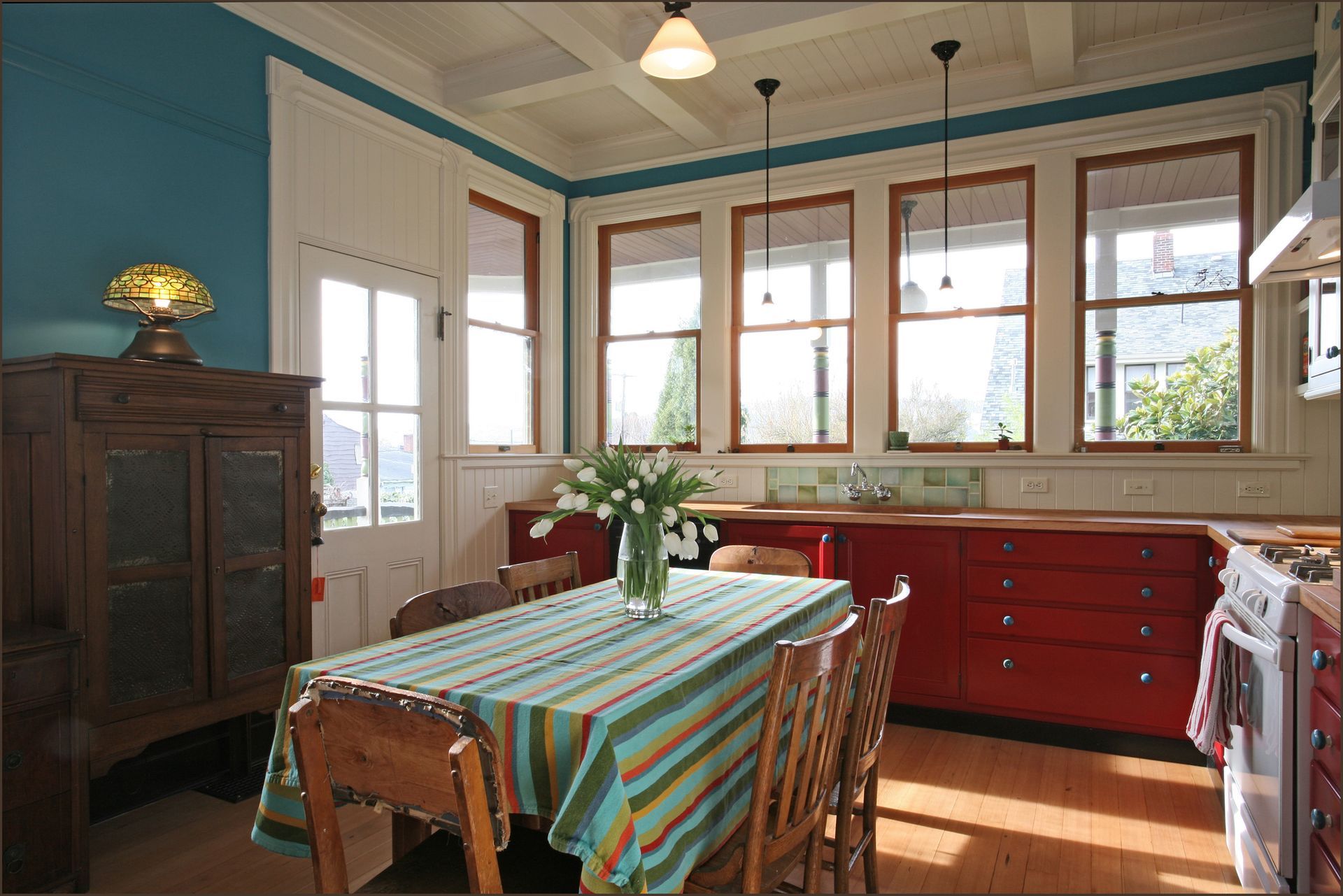 Bright dining room with striped tablecloth, red sideboard, and large windows with daylight