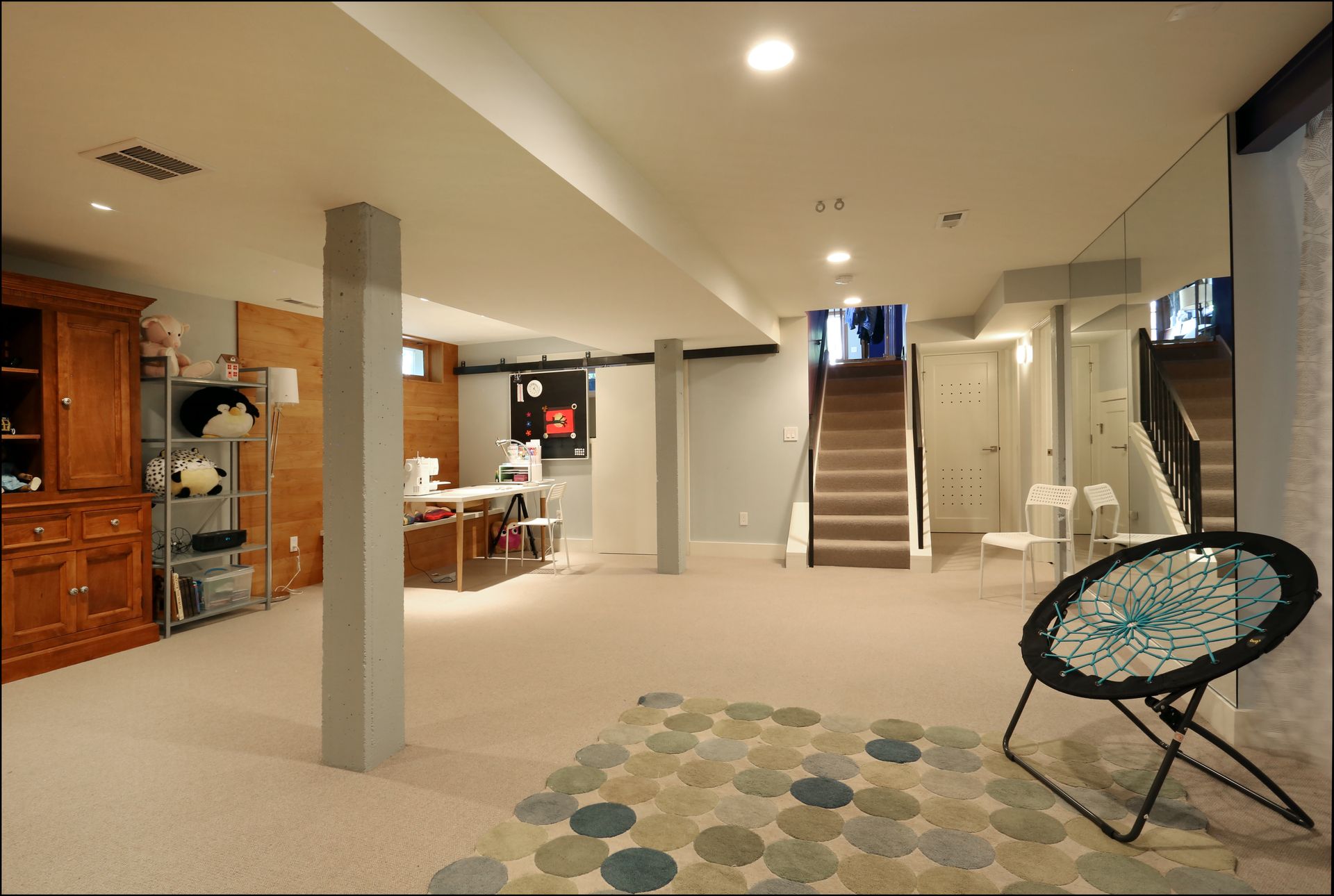 Spacious beige basement with stairs, kitchen area, column, patterned rug, and black chair