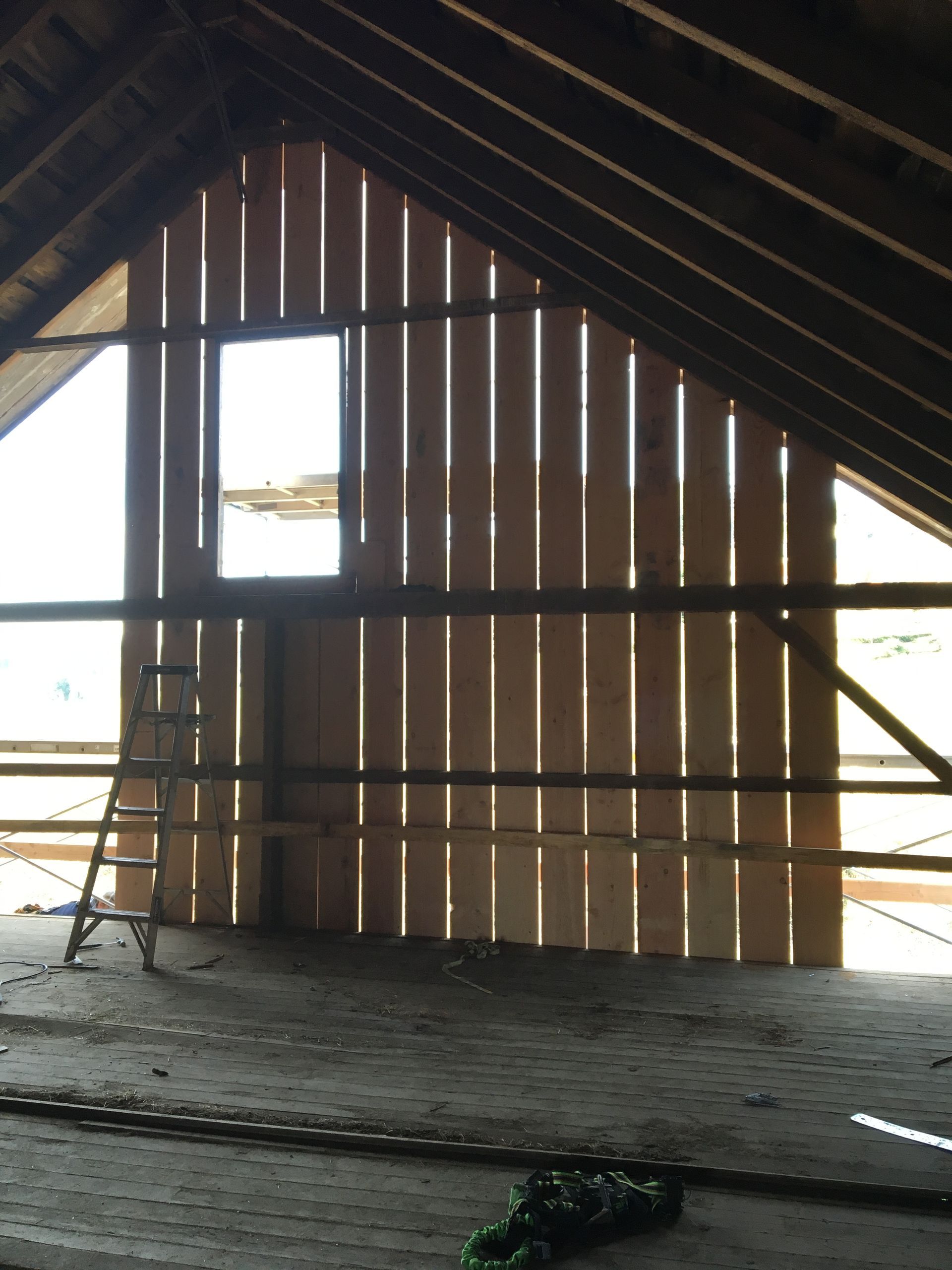 Empty attic with exposed wooden beams, slatted wall, and a ladder near a bright window opening.