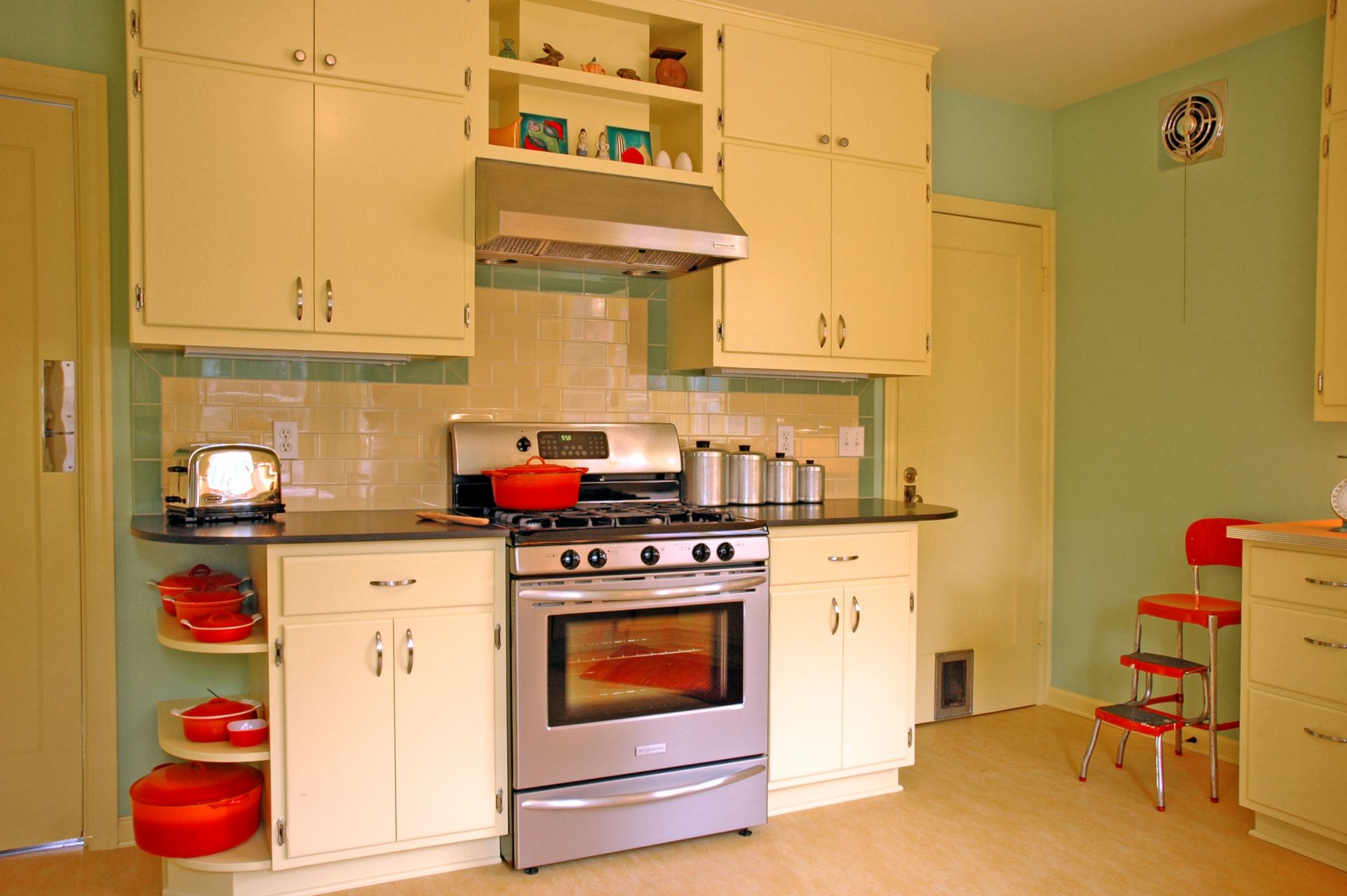 Small mint-green kitchen with white cabinets, stainless stove, and red cookware on counters and shelves