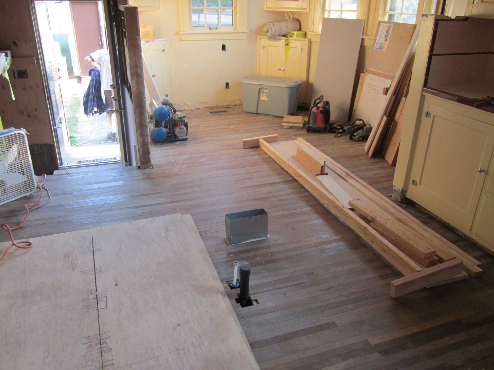 Renovation in progress in a room with tools, lumber, and plywood on the floor near cabinets and windows.