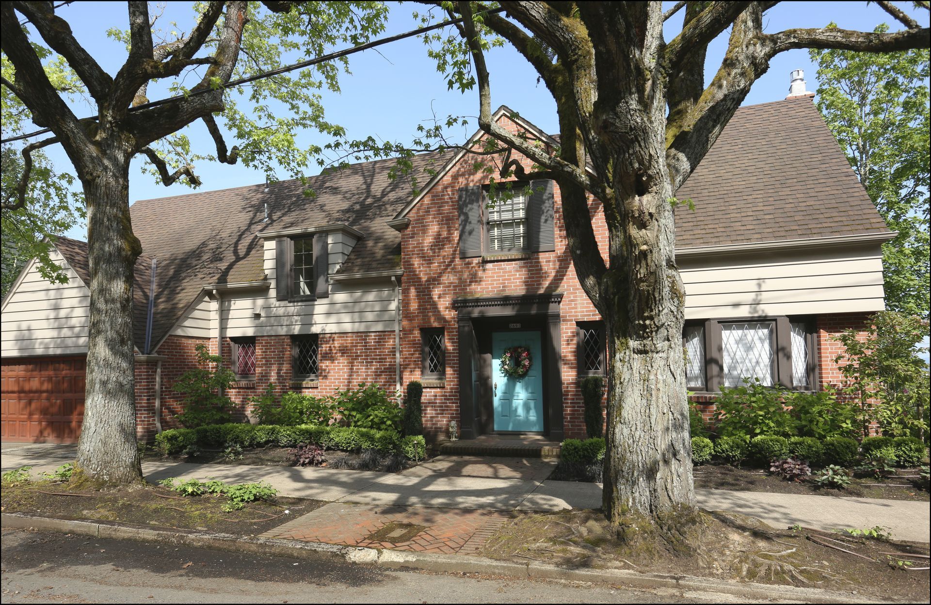 Brick house with steep roof, teal front door, and large trees along the sidewalk