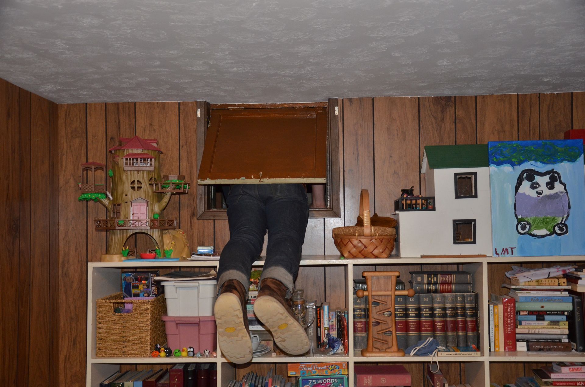 Person climbing into a wall opening above shelves in a wood-paneled room, feet hanging out.