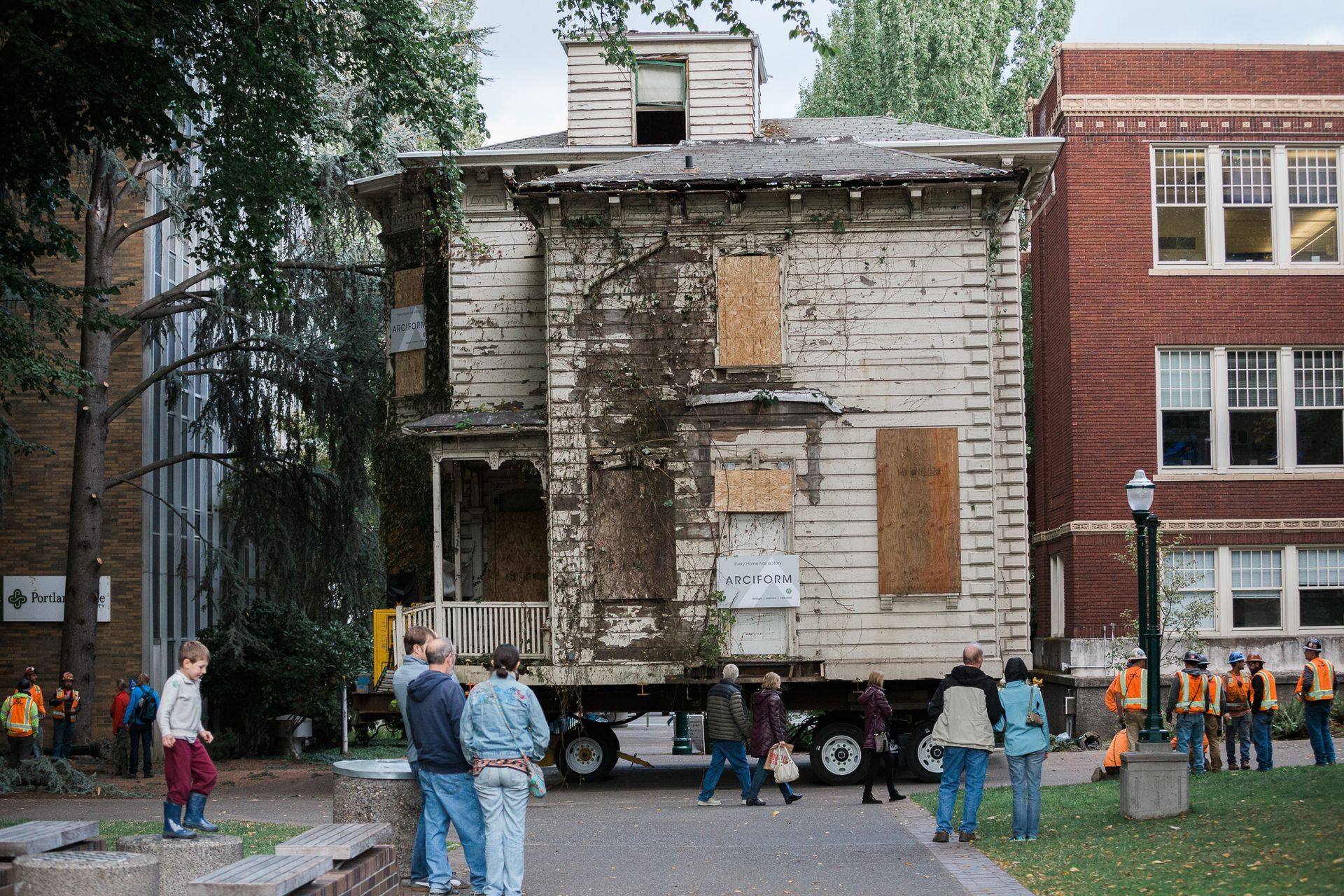 Old house facade being relocated on a truck, with people watching in a grassy courtyard.