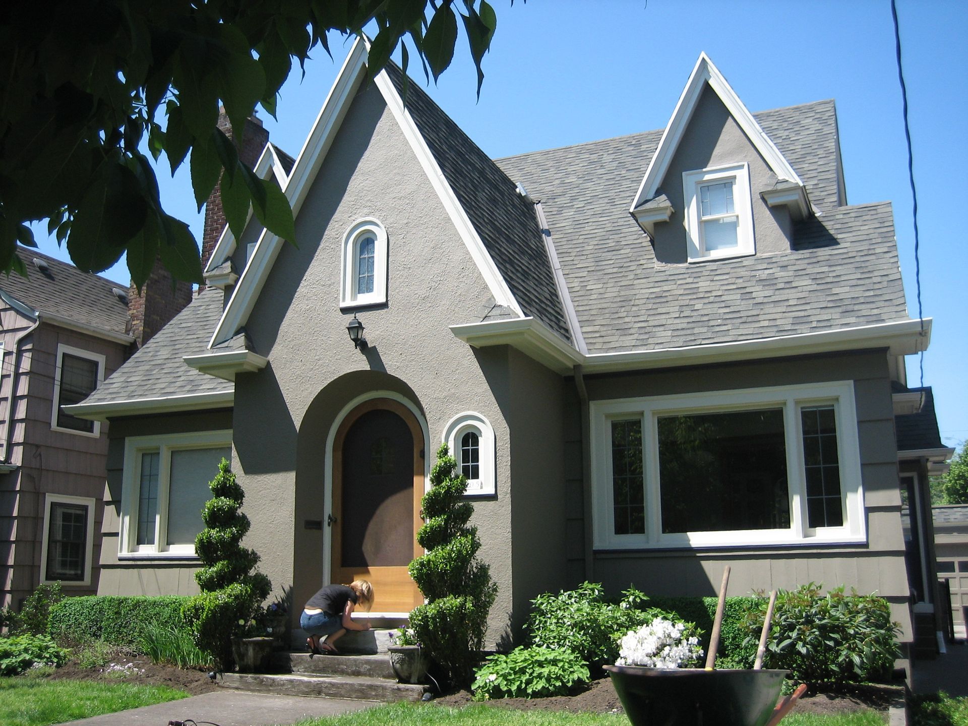 Gray stone house with steep gables, arched front door, and white-trimmed windows, framed by a sunny garden.