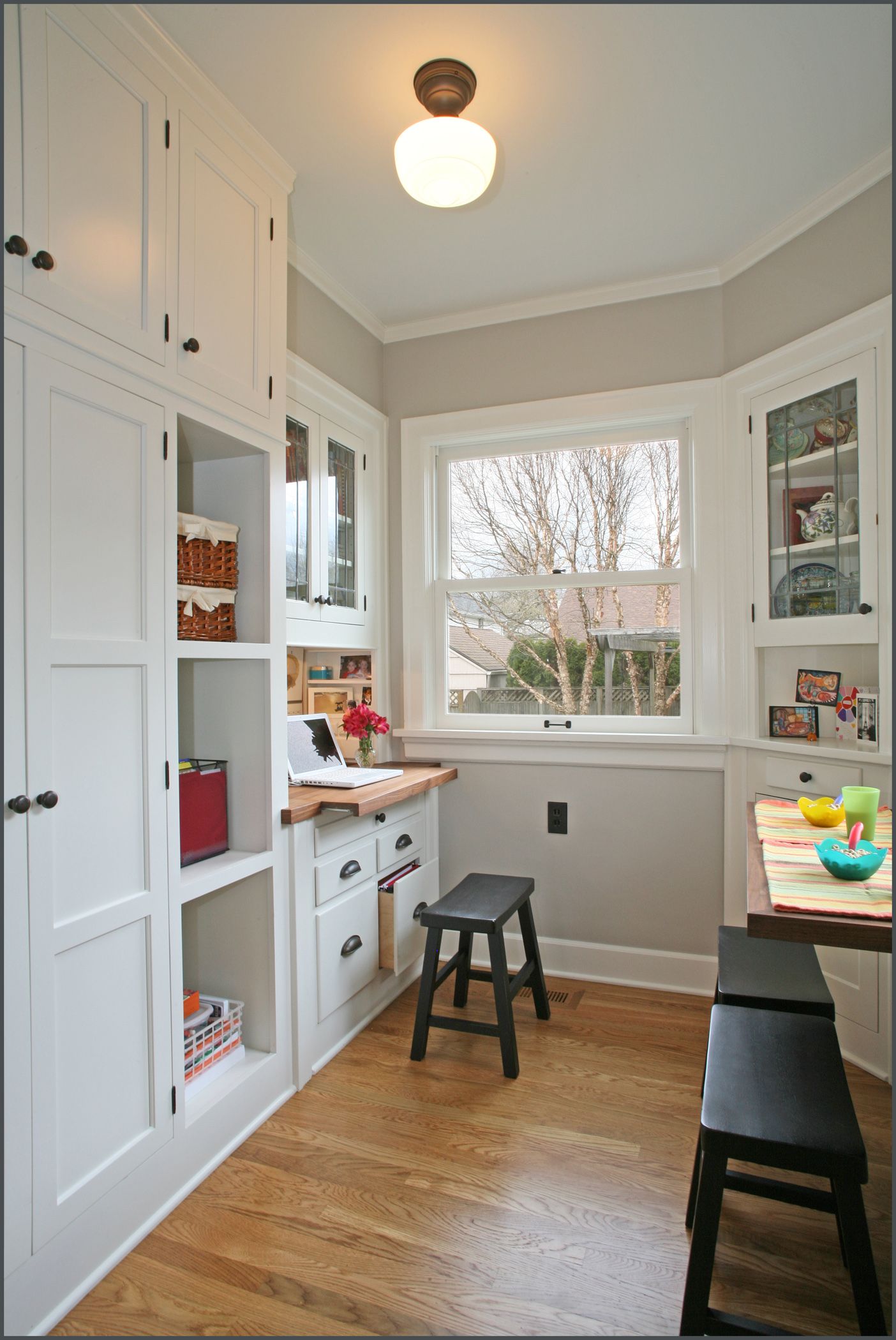 Narrow white kitchen with built-in cabinets, bay window, wooden floor, and a small stool.
