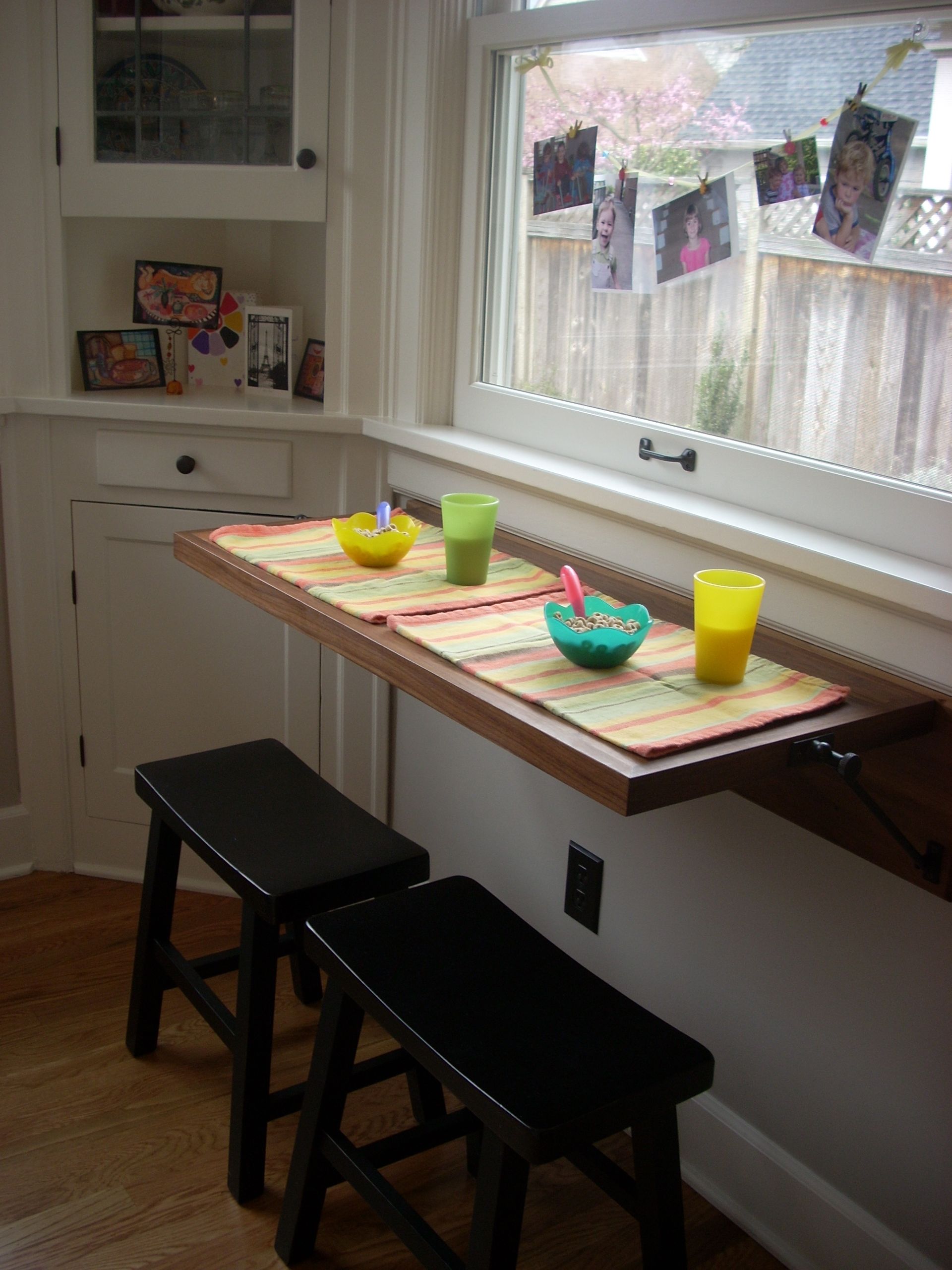 Small kitchen nook with two black stools, a wooden counter, and colorful cups by a window with hanging photos.