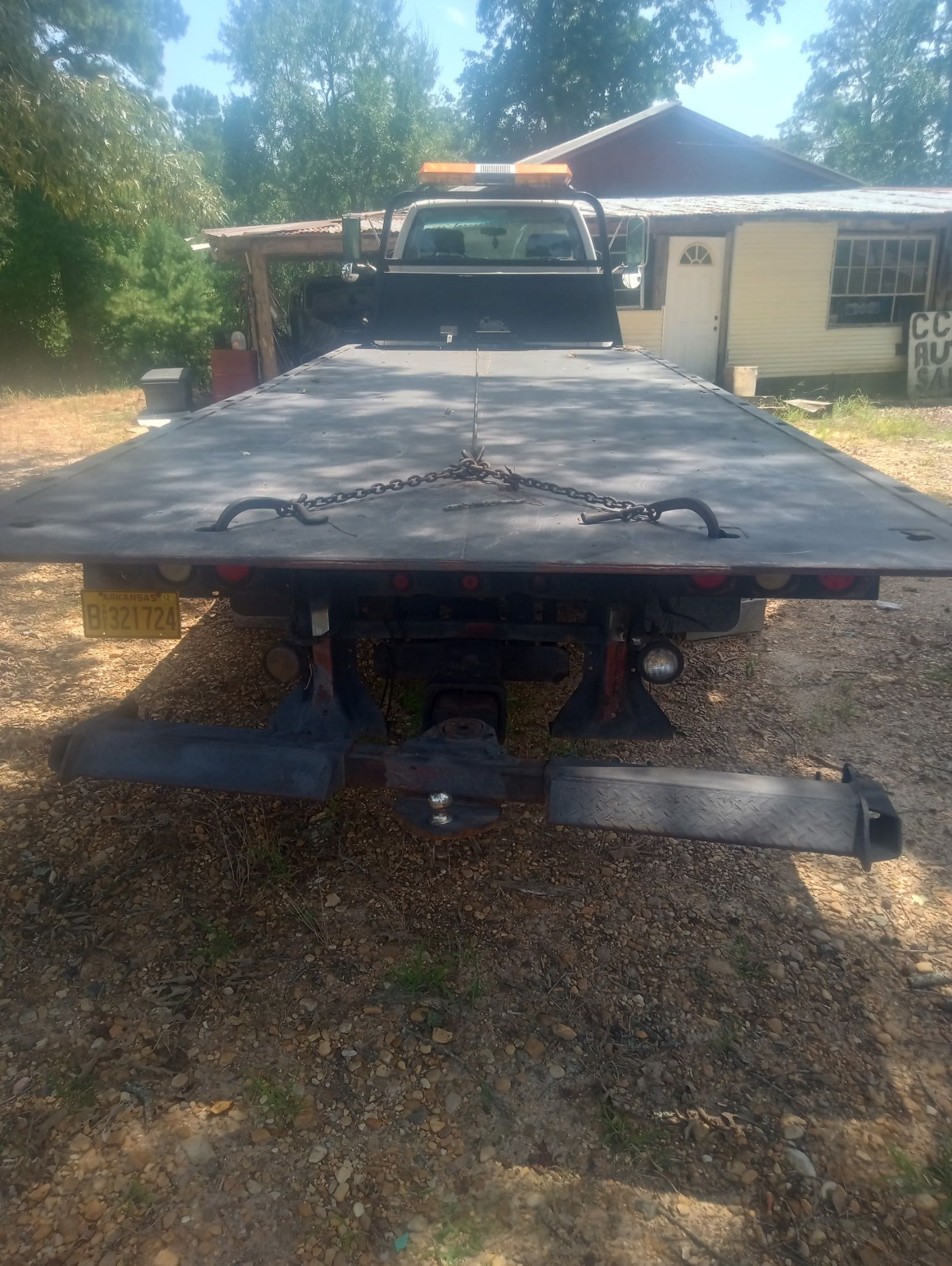 Flatbed tow truck parked on gravel driveway beside a house and trees, front view with raised cab light bar.