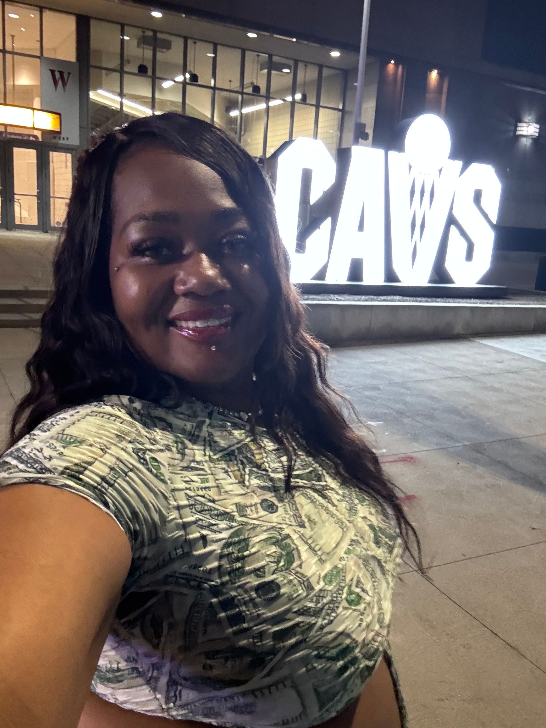 Woman smiling in a patterned top taking a selfie at night in front of a bright CAVS sign.