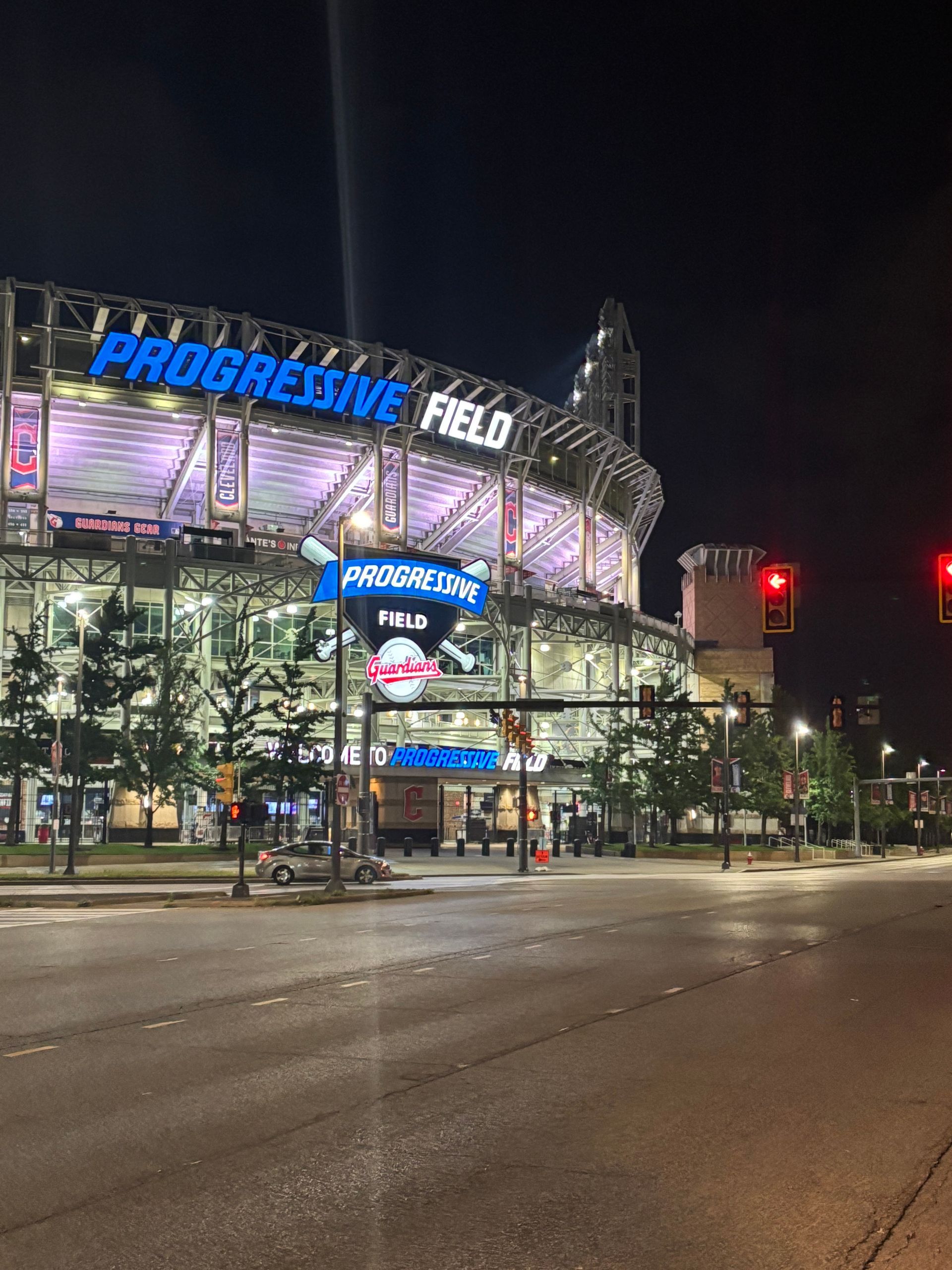 Progressive Field lit up at night with streetlights and a nearly empty road in front.