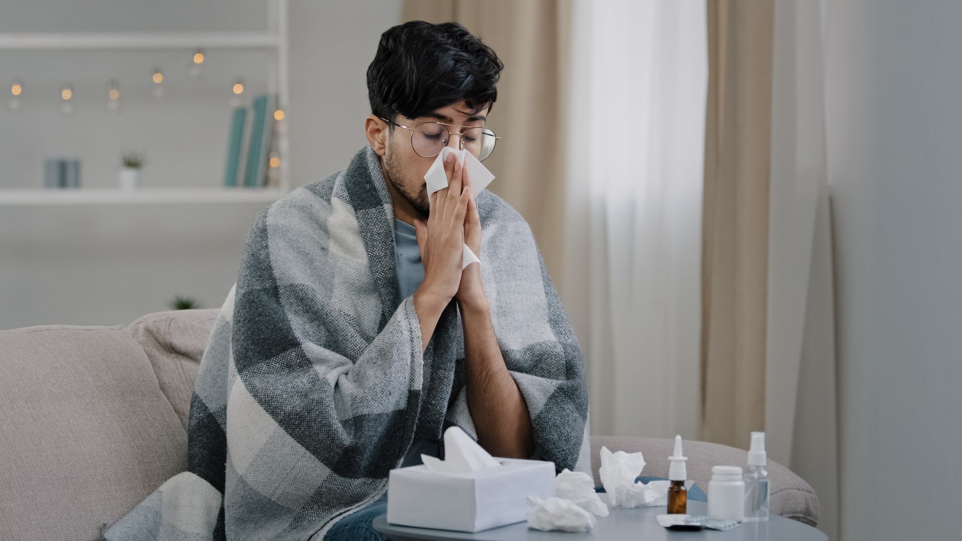 A man is sitting on a couch blowing his nose into a napkin.
