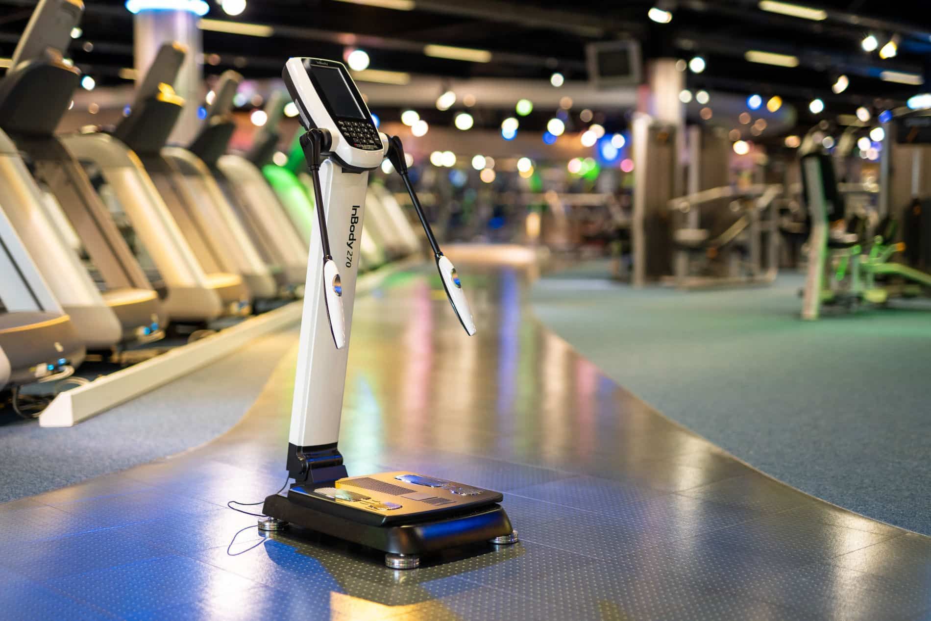 Body composition analyzer in a gym, with treadmills and equipment in the background.