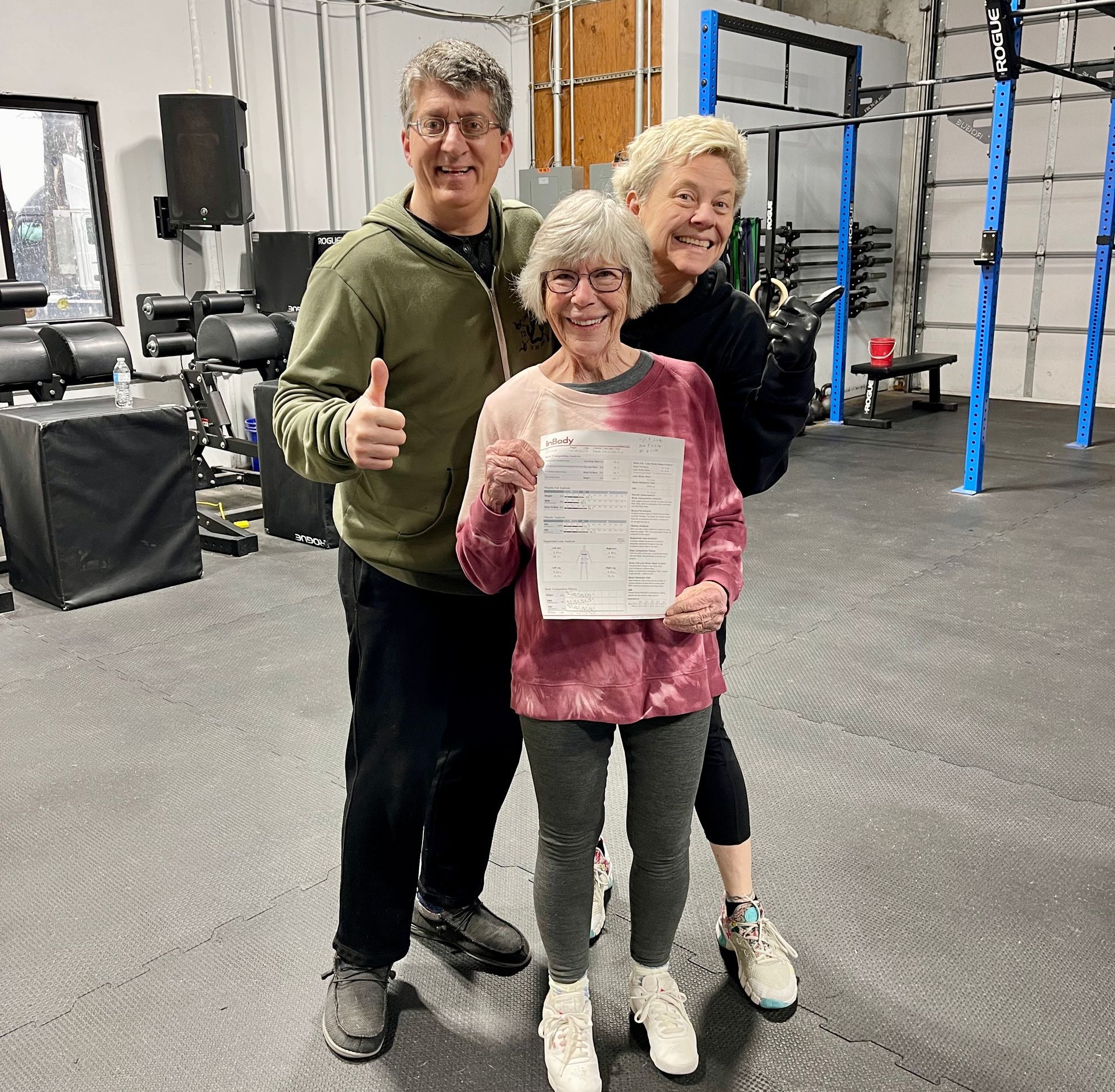 Three people in a gym. A woman holds a paper, smiling. The others give thumbs up.
