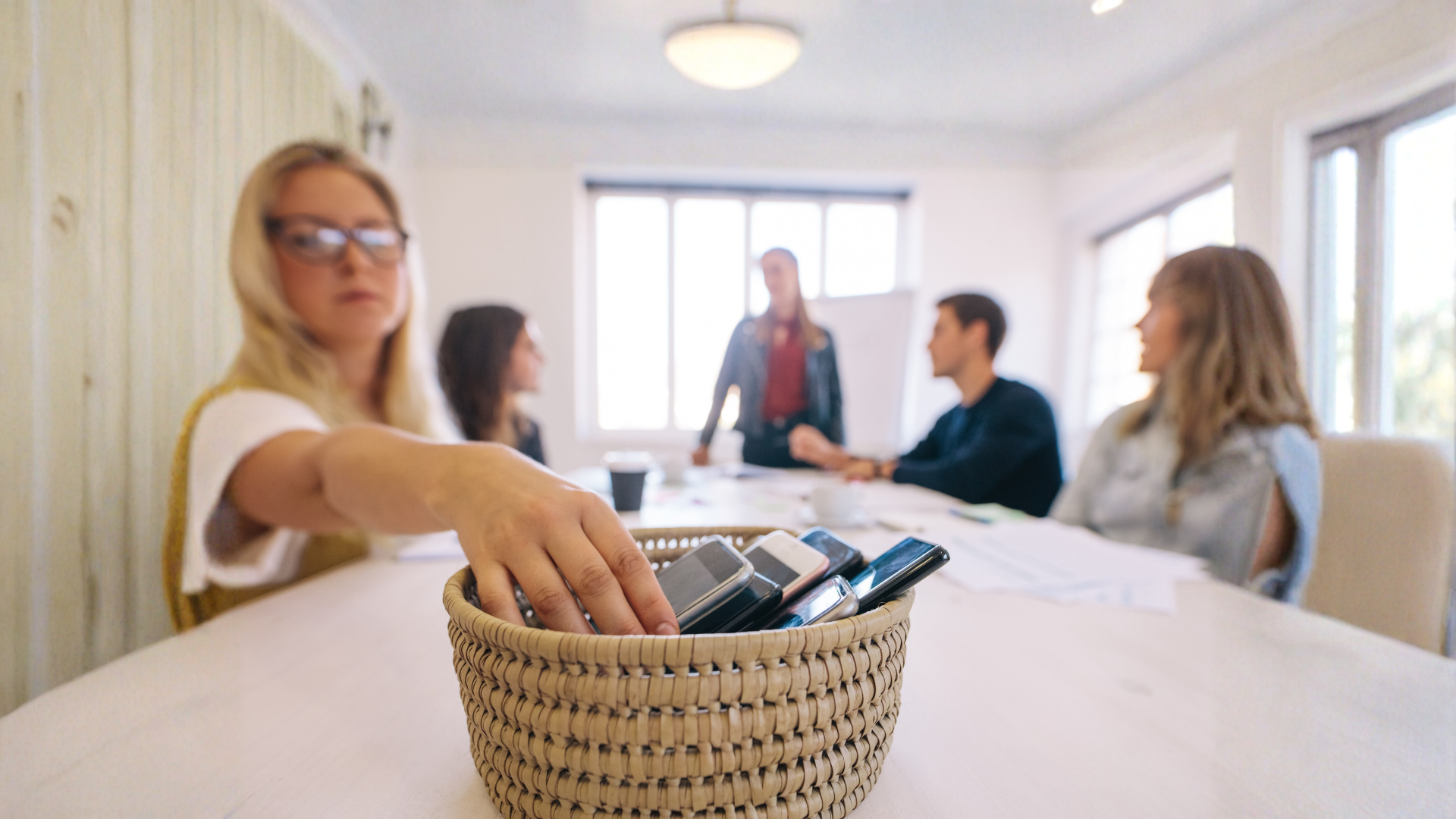 Woman reaches into a basket of pens and markers in a bright meeting room while others talk in the background