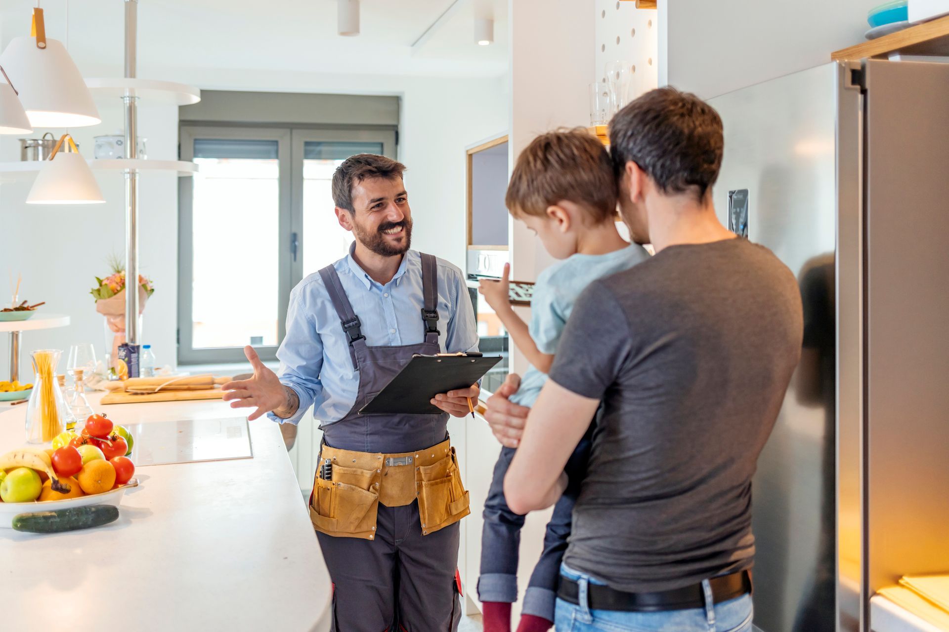 A man is holding a child while talking to a plumber in a kitchen.