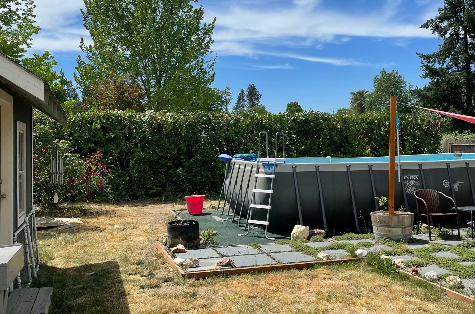Backyard with above-ground pool, ladder, red bucket, patio stones, and trees under a blue sky