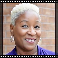 A woman with short white hair and hoop earrings is smiling in front of a brick wall.