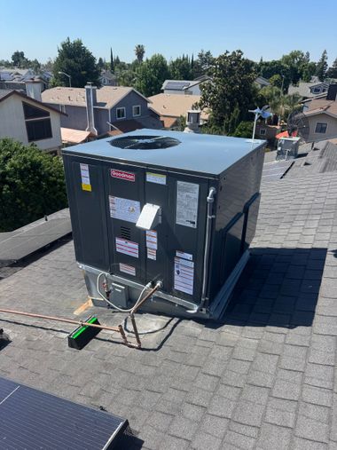 Rooftop HVAC unit with attached cables and labels on a shingled roof in a residential neighborhood