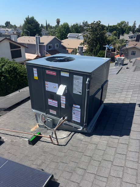 Rooftop HVAC unit with attached cables and labels on a shingled roof in a residential neighborhood