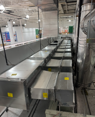 Rows of silver HVAC ducts and vents in an industrial ceiling space, with black piping and metal beams.