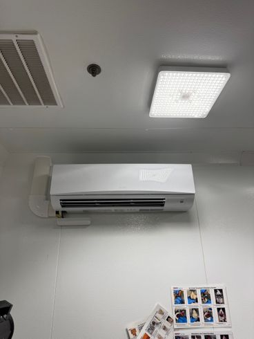 Wall-mounted white air conditioner beneath a ceiling vent and light, with small photos on the wall below.
