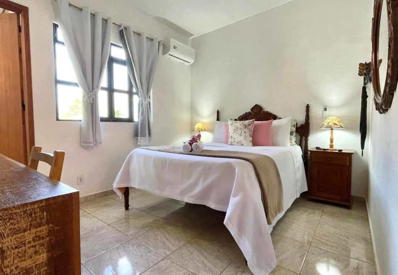 Quarto iluminado com cama arrumada, cortinas brancas, mesas de cabeceira e piso de azulejo.