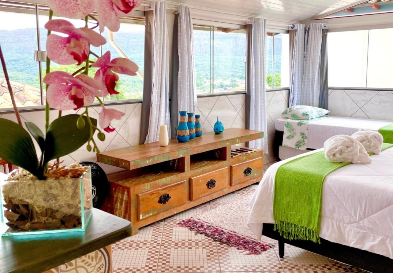 Quarto iluminado com janelas amplas, cômoda de madeira, cobertor verde na cama e orquídea em vaso em primeiro plano.